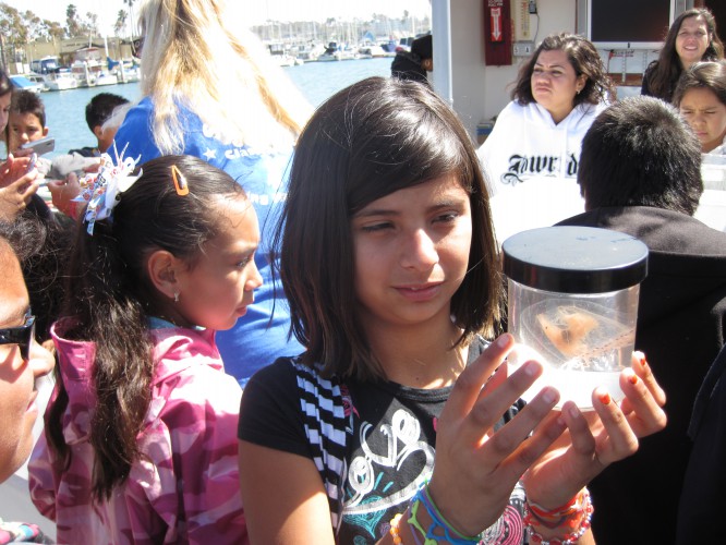 A girl holds a clear container with an aquatic specimen while standing on a boat with other people. Water and docked boats are visible in the background.