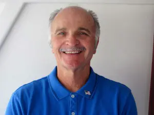 A smiling man with thinning gray hair and a mustache is wearing a blue collared shirt with a small flag pin on it. He is standing against a plain light-colored background.